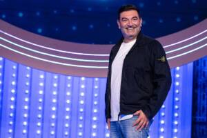 Max Giusti di profilo sorride a favore di camera con maglietta bianca e giacca nera