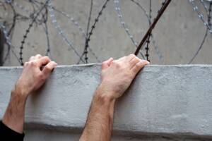 Mani aggrappate al bordo di un muro sormontato da filo spinato, durante un tentativo di fuga.
