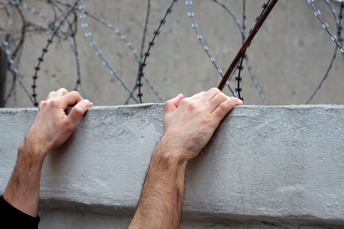 Mani aggrappate al bordo di un muro sormontato da filo spinato, durante un tentativo di fuga.