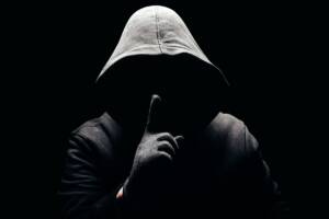 Uomo incappucciato nell'oscurità che fa il gesto del silenzio con l'indice.