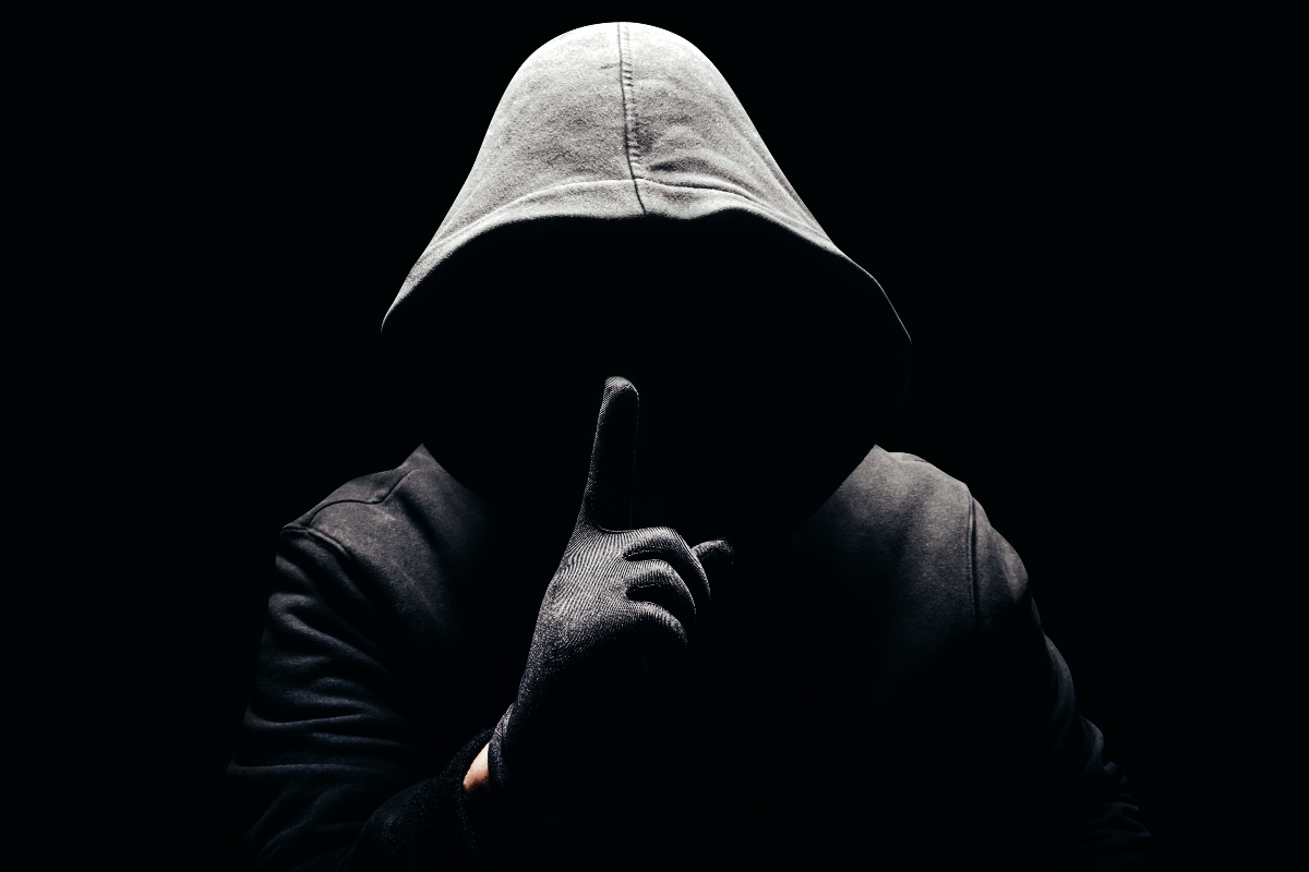 Uomo incappucciato nell'oscurità che fa il gesto del silenzio con l'indice.